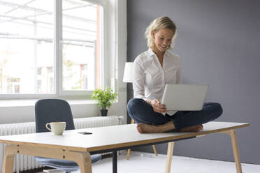 Lächelnde junge Geschäftsfrau sitzt auf dem Schreibtisch im Büro und benutzt einen Laptop - MOEF02691