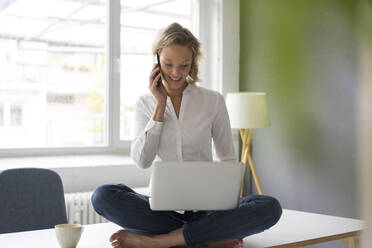 Lächelnde junge Geschäftsfrau sitzt auf dem Schreibtisch im Büro und benutzt Laptop und Mobiltelefon - MOEF02692