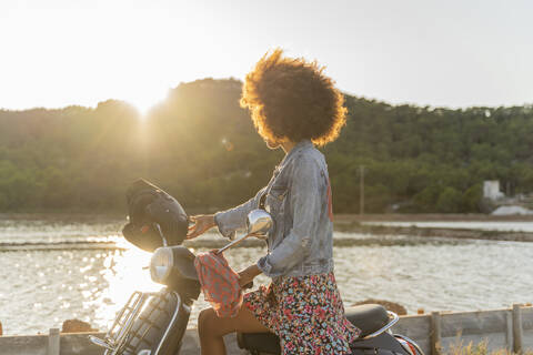 Junge Frau auf einem Motorroller sitzend bei Sonnenuntergang, Ibiza, lizenzfreies Stockfoto