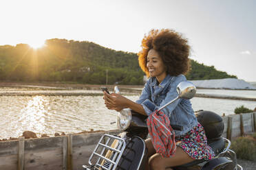Junge Frau sitzt auf einem Motorroller und benutzt ihr Smartphone bei Sonnenuntergang, Ibiza - AFVF04360