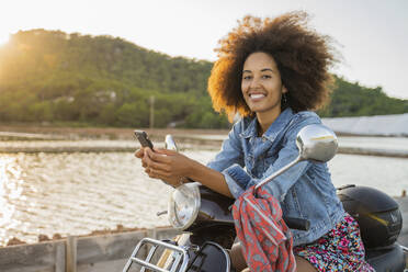 Junge Frau sitzt auf einem Motorroller und benutzt ihr Smartphone bei Sonnenuntergang, Ibiza - AFVF04361