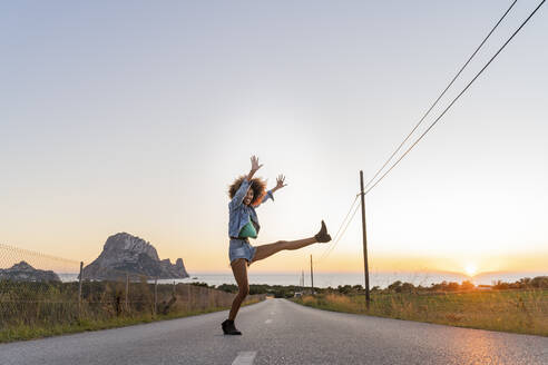 Junge Frau steht auf der Straße und hebt ihr Bein bei Sonnenuntergang, Ibiza - AFVF04374