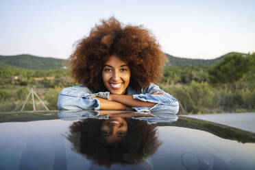 Young woman during road trip, leaning on car roof and looking at camera - AFVF04390