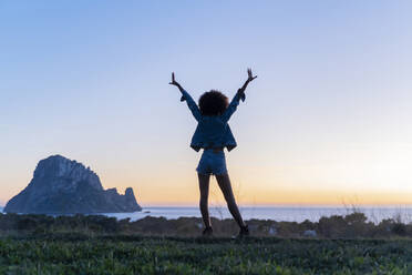 Rückansicht einer jungen Frau, die bei Sonnenuntergang mit erhobenen Armen auf einem Aussichtspunkt steht, Ibiza - AFVF04404