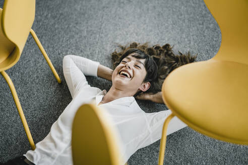 Laughing businesswoman laying in an office on the floor between chairs - KNSF06867