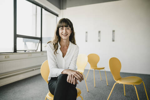 Lächelnde Geschäftsfrau sitzt auf einem Stuhl in einem leeren Büro, lizenzfreies Stockfoto