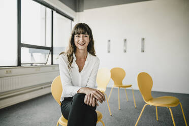Smiling businesswoman sitting on a chair in an empty office - KNSF06868