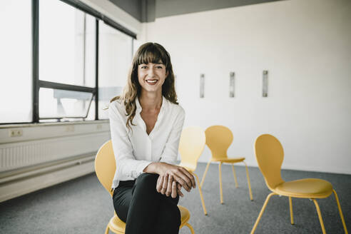 Smiling businesswoman sitting on a chair in an empty office - KNSF06868