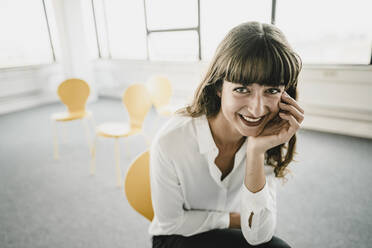 Smiling businesswoman sitting on a chair in an empty office - KNSF06870