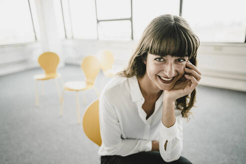 Lächelnde Geschäftsfrau sitzt auf einem Stuhl in einem leeren Büro - KNSF06870