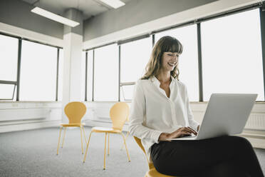 Lächelnde Geschäftsfrau, die auf einem Stuhl in einem leeren Büro sitzt und einen Laptop benutzt - KNSF06871
