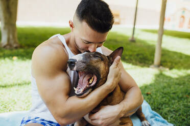 Muscular man sitting on blanket on a meadow hugging his dog - MIMFF00001