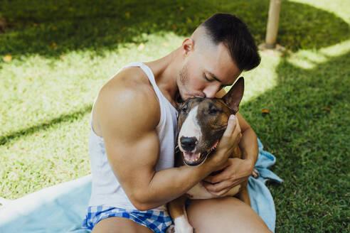 Muscular man sitting on blanket on a meadow hugging his dog - MIMFF00002