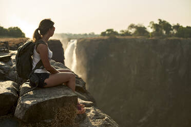 Woman sitting on the top of a rock enjoying the Victoria Falls at sunset, Zimbabwe - VEGF01052