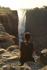 Woman sitting on the top of a rock enjoying the Victoria Falls at sunset, Zimbabwe - VEGF01056