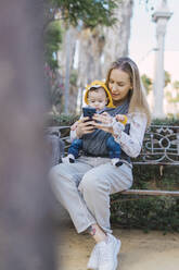 Mutter ruht sich mit ihrem kleinen Jungen auf einer Parkbank aus und benutzt ihr Smartphone - ERRF02236