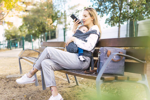 Mutter ruht sich mit ihrem kleinen Jungen auf einer Parkbank aus und benutzt ihr Smartphone - ERRF02243
