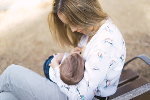 Mother breastfeeding baby boy on a park bench - ERRF02248