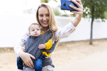 Glückliche Mutter mit Baby in einem Tragetuch macht ein Selfie - ERRF02282