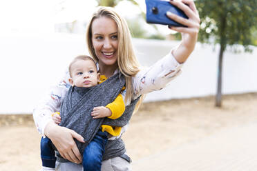 Happy mother with baby boy in a sling taking a selfie - ERRF02282