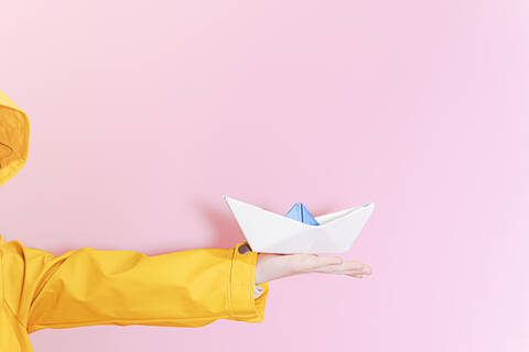 Kleines Mädchen, das ein Ölzeug trägt und ein Papierboot auf dem ausgestreckten Arm hält, lizenzfreies Stockfoto