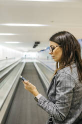 Young businesswoman using smartphone on moving walkway - JRFF03901