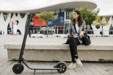 Junge Geschäftsfrau mit Smartphone neben einem E-Scooter in der Stadt - JRFF03906