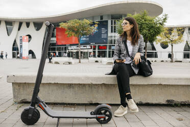 Young businesswoman holding smartphone next to an e-scooter in the city - JRFF03906