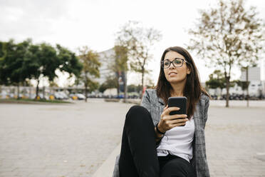Junge Geschäftsfrau mit Smartphone, auf einer Bank in der Stadt sitzend - JRFF03910