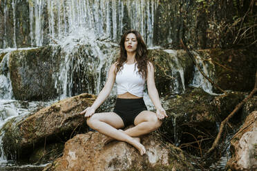 Junge Frau übt Yoga an einem Wasserfall, auf einem Felsen sitzend - MIMFF00011