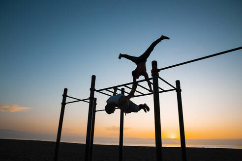 Zwei junge Männer, die bei Sonnenaufgang in einer Turnhalle im Freien Gymnastik machen - MIMFF00019