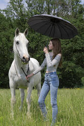 Teenage girl standing on a meadow with horse and umbrella - PSTF00507