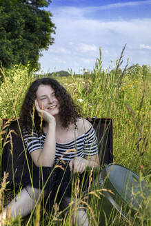 Porträt einer glücklichen jungen Frau, die im Koffer auf einer Wiese sitzt und in die Ferne schaut - PSTF00538