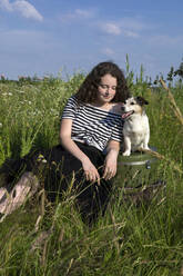 Portrait of young woman sitting on a meadow with suitcases and her little dog - PSTF00555