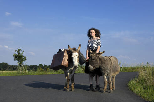 Junge Frau steht auf einer Landstraße mit Eseln, die Gepäck tragen - PSTF00559
