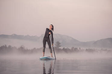Woman stand up paddling on lake Kirchsee at morning mist, Bad Toelz, Bavaria, Germany - WFF00197