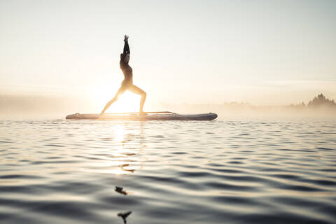 Frau beim morgendlichen Yoga mit dem Paddle Board auf dem Kirchsee, Bad Tölz, Bayern, Deutschland, lizenzfreies Stockfoto