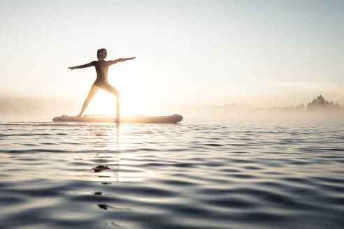 Woman practicing paddle board yoga on lake Kirchsee in the morning, Bad Toelz, Bavaria, Germany - WFF00204