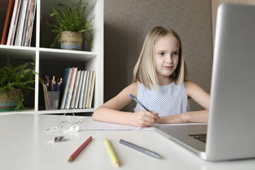 Girl sitting at table at home doing homework and using laptop - EYAF00747