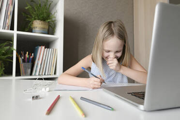 Laughing girl sitting at table at home doing homework and using laptop - EYAF00748