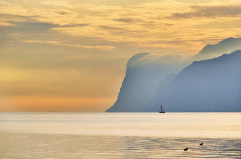 Italien, Trentino, Nago-Torbole, Silhouette eines Segelboots in der Nähe der Küstenklippen des Gardasees bei stimmungsvoller Morgendämmerung - MRF02310