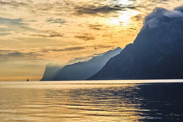 Italien, Trentino, Nago-Torbole, Silhouette eines Segelboots in der Nähe der Küstenklippen des Gardasees bei stimmungsvollem Sonnenaufgang - MRF02313