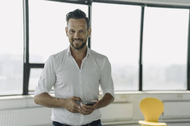 Porträt eines selbstbewussten reifen Geschäftsmannes mit Mobiltelefon in einem leeren Büro - KNSF06881