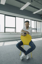 Porträt eines selbstbewussten reifen Geschäftsmannes, der auf einem gelben Stuhl in einem leeren Büro sitzt - KNSF06882