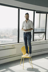 Glücklicher reifer Geschäftsmann steht auf gelbem Stuhl in leerem Büro - KNSF06884