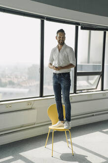 Happy mature businessman standing on yellow chair in empty office - KNSF06884