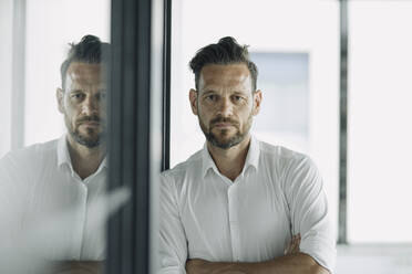 Portrait of serious mature businessman leaning against glass wall in office - KNSF06904