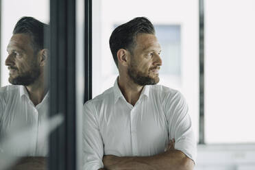 Mature businessman leaning against glass wall in office looking sideways - KNSF06905