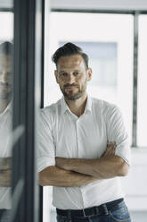 Portrait of confident mature businessman leaning against glass wall in office - KNSF06906