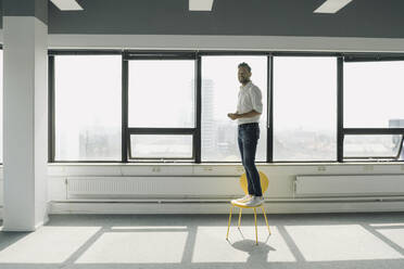 Happy mature businessman standing on yellow chair in empty office - KNSF06914